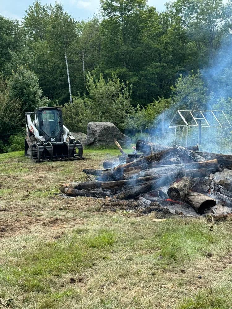 Bobcat loader beside a smoldering log pile in a wooded area, with greenery in the background.