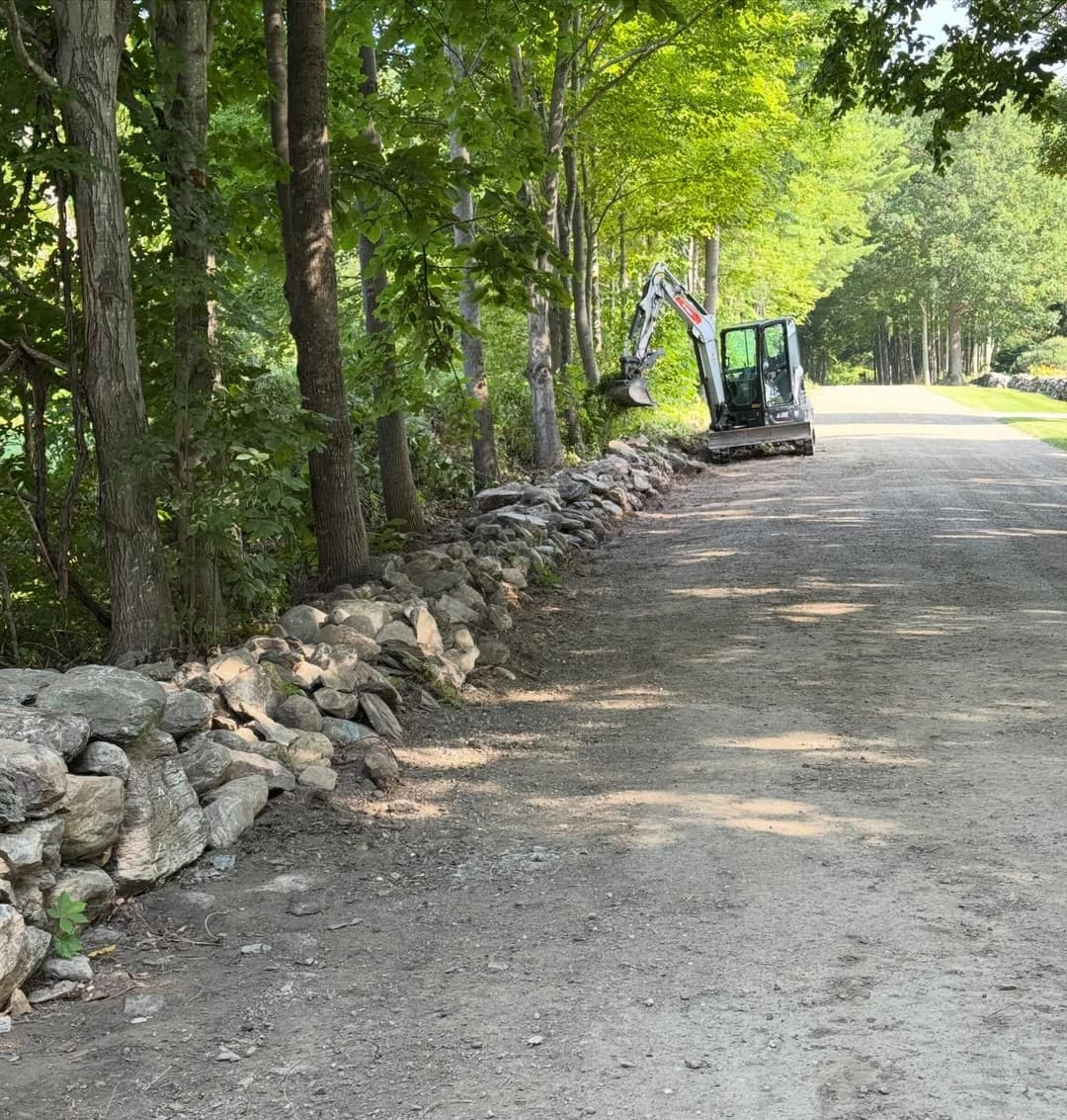 Excavator working beside a stoned path in a wooded area on a sunny day.