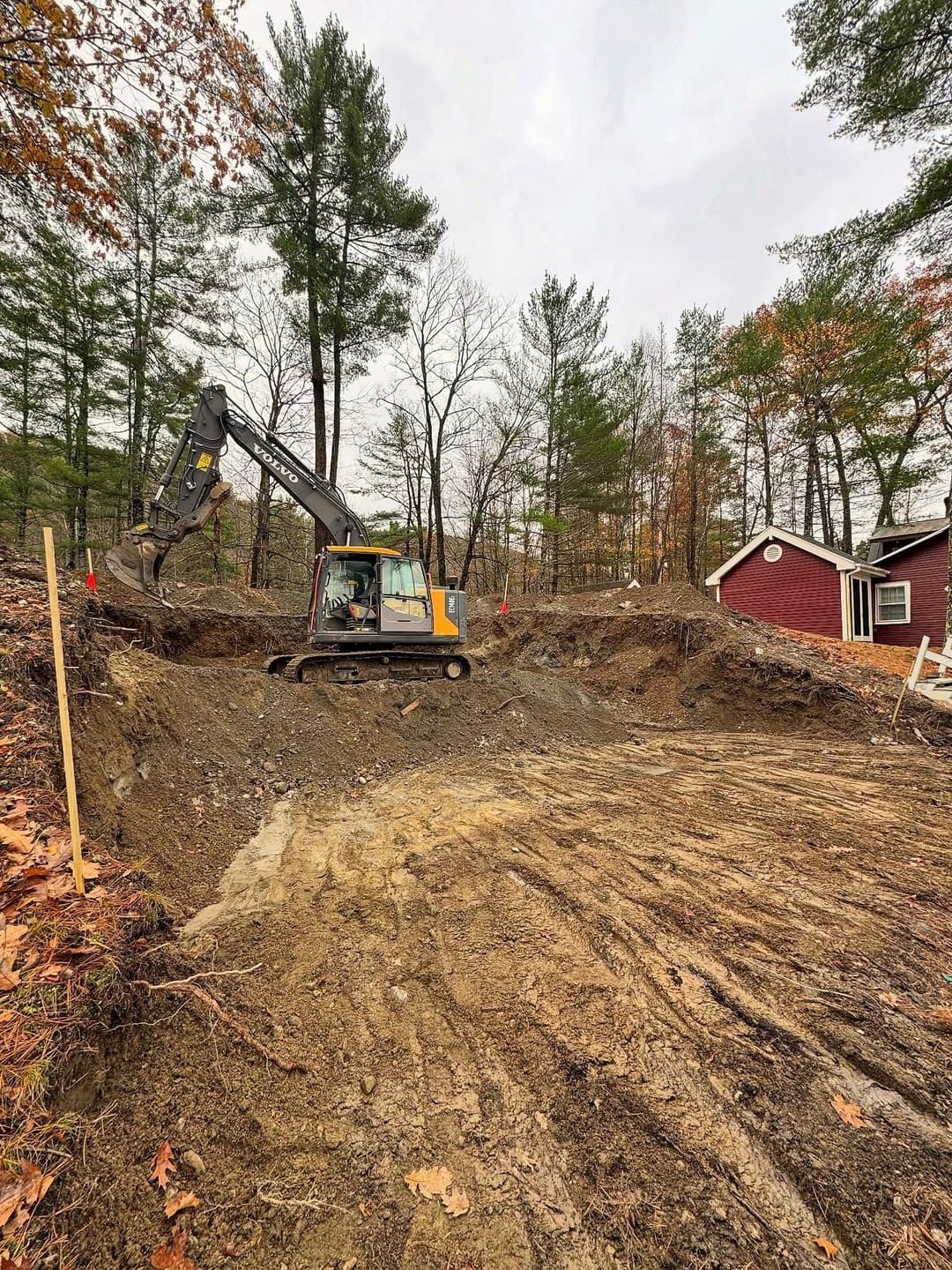 Excavator digging foundation on residential construction site surrounded by trees.