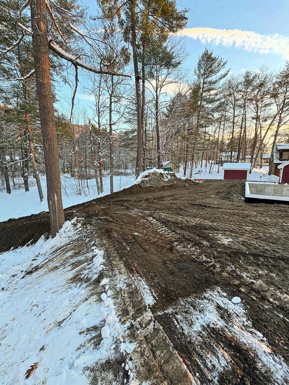 Snow-covered construction site with freshly graded land among pine trees at sunset.