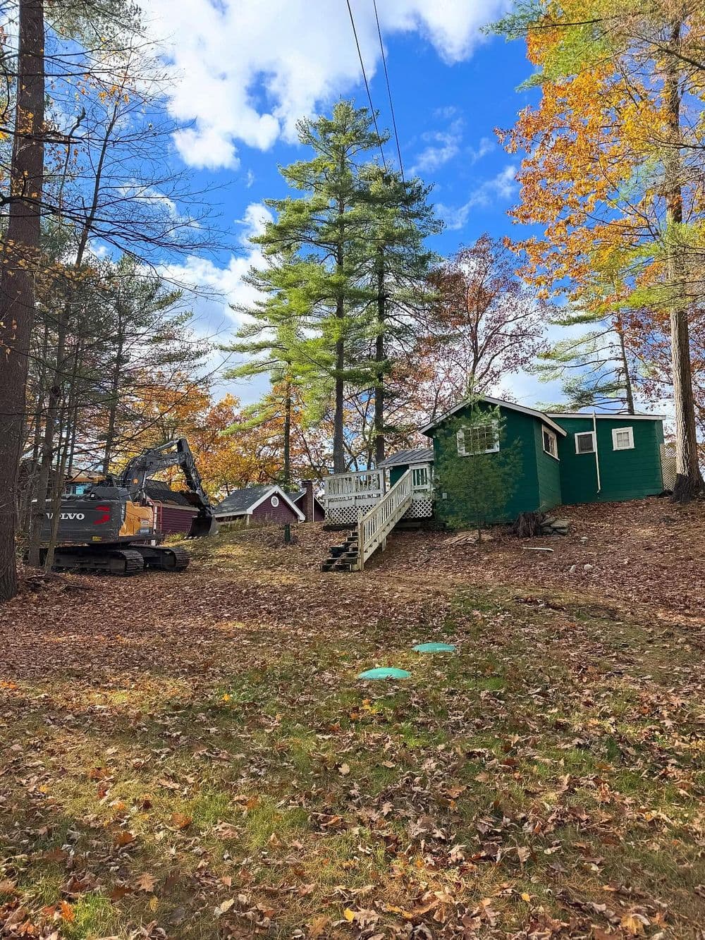 Green cabin surrounded by autumn foliage, with an excavator nearby in a wooded area.