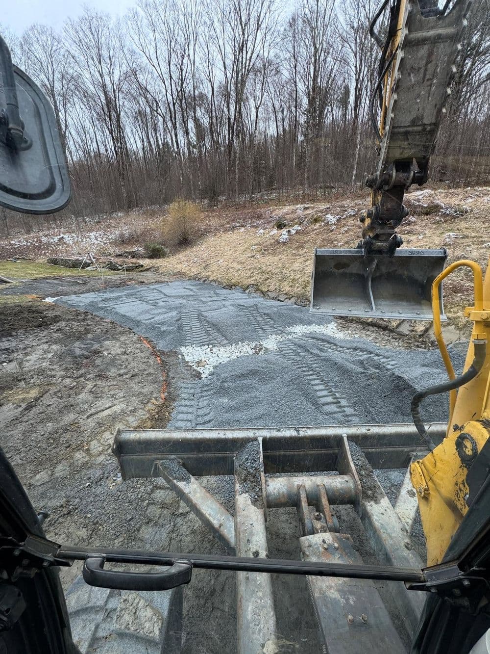 Excavator smoothing gravel path in a forested area during winter.