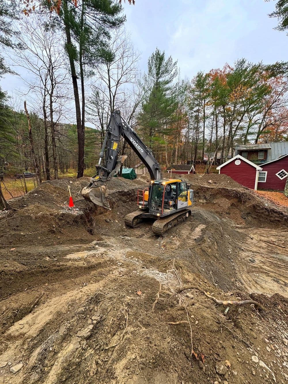 Excavator digging in a construction site surrounded by trees and a red building.