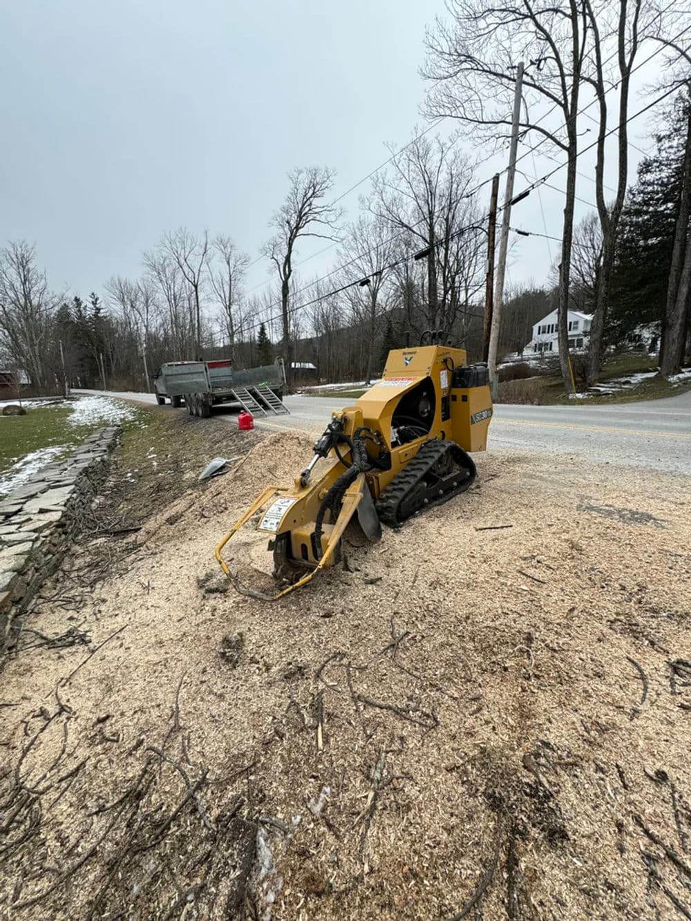 Stump grinder on a rural road, surrounded by wood chips and trees in winter setting.
