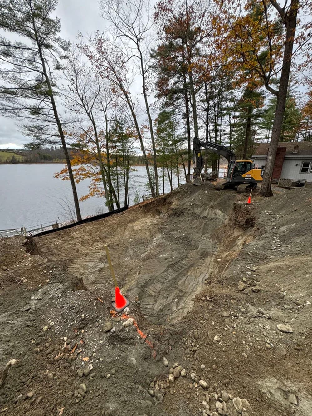 Construction site near a lake with heavy machinery, surrounded by trees and autumn foliage.